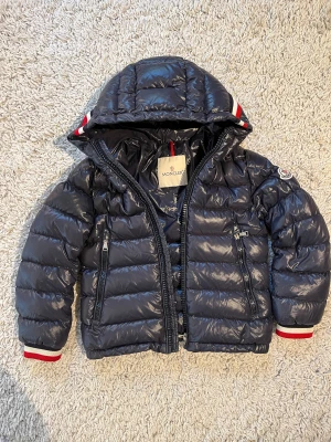 Moncler dunjacka för barn - Snygg mörkblå dunjacka från Moncler med glansig finish, huva och dragkedjor framtill. Den passar barn i 8 års åldern 130cm. Jackan har Moncler-logga på ärmen och coola rödvita detaljer vid muddar och huva. Fylld med vitt gåsdun för extra värme och stilpoäng. Den är såklart äkta som ni kan se på sista bilden! Skriv vid minsta fundering.