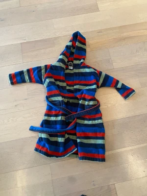 Randig badrock med huva från Åhléns - Supermysig badrock från Åhléns i storlek 98-104, perfekt för barn 3-4 år. Badrocken har huva, knytskärp i midjan och coola breda ränder i blått, rött, grönt och mörkblått. Tillverkad i mjuk frotté som torkar snabbt efter badet.