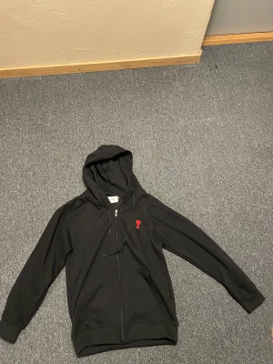 Svart hoodie från Ami med röd logga - Svart hoodie från Ami Alexandre Mattiussi med dragkedja, huva och snörning. På bröstet sitter Ami:s ikoniska röda hjärta-broderi. Tröjan har kängurufickor och är tillverkad i mjuk bomull. Perfekt för en chill och stilren look. OBS den är liten i storlek, den är medium men den passar som small
