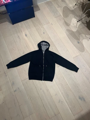 Svart  hoodie med mönstrad insida - Svart hoodie med hel dragkedja och snörning i huvan. Insidan har ett stilrent svartvitt mönster som ger extra lyxig känsla. Tröjan har långärmade ärmar och normal passform, perfekt för dig som gillar exklusiva streetwear-vibbar.