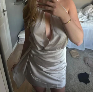 Champagnefärgad omlottklänning i satin - Snygg kort klänning i champagnefärgad satin med omlott-detalj och djup v-ringning. Klänningen har halterneck och knyt i sidan som ger en cool draperad effekt. Perfekt för dig som vill sticka ut med en elegant och trendig look.