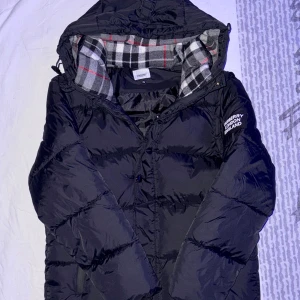 Svart pufferjacka från Burberry - Säljer en svart pufferjacka från Burberry med huva och logotyp på ärmen. Jackan har en klassisk, puffig design med knappar och dragkedja framtill. Perfekt för kalla dagar och har en riktigt snygg streetstil.