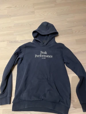 Mörkblå hoodie Peak Performance - Snygg mörkblå hoodie från Peak Performance med vit logotyp på bröstet. Tröjan har huva, lång ärm och en klassisk känguruficka framtill. Mjuk bomullskvalitet och ribbade muddar vid ärmslut och nederkant. Perfekt för chill eller streetwear. Vid funderingar skriv. Pris kan diskuteras kom med förskag.
