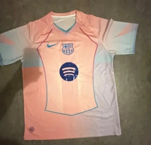 Barça kortärmad fotbollströja Nike - Säljer en Barcelona fotbollströja från Nike i ljusrosa och ljusblå toner med klubbmärke och Spotify-logga på bröstet. Tröjan har V-ringning med turkos kant och är tillverkad i lätt, ventilerande polyester. Perfekt för dig som vill sticka ut på planen.