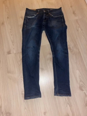 Dondup George blå jeans slim fit - Snygga blå jeans från Dondup, modellen George. Slim fit med klassisk femficksdesign och låg midja. Jeansen har slitningar vid fickorna och snygga detaljer i sömmarna. Perfekt för dig som gillar en modern och stilren look.