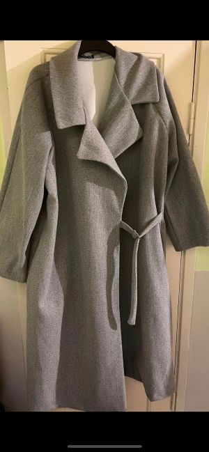 Grå kappa med bälte och bred krage - Stilren grå kappa med bred krage och knytbälte i midjan. Långärmad och rak modell som ger en clean och modern look. Perfekt för dig som gillar minimalistisk stil och vill ha en jacka som funkar till många outfits.