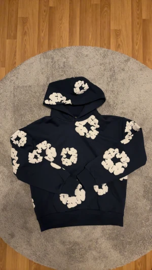 Mörkblå Denim tears Hoodie  - Säljer en mörkblå hoodie från Denim Tears i storlek L med unikt vitt blommigt mönster över hela tröjan. Hoodien har huva och långa ärmar, tillverkad i mjuk bomull. Perfekt för dig som gillar streetwear och vill sticka ut med cool design.