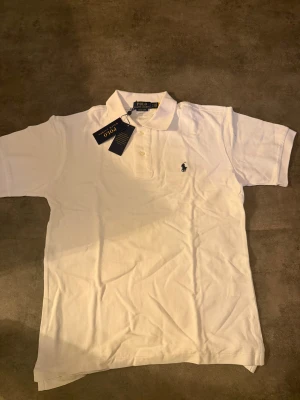 Vit pikétröja Polo Ralph Lauren M - Vit pikétröja från Polo Ralph Lauren i modellen Custom Slim Fit. Klassisk krage, kort ärm och broderad marinblå logga på bröstet. Tröjan är i bomull och har knappar framtill. Perfekt för en clean och stilren look.