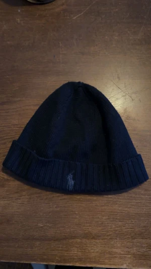 Mörkblå stickad mössa från Polo Ralph Lauren - Snygg mörkblå stickad mössa från Polo Ralph Lauren med klassisk ribbad kant och broderad logga framtill. Perfekt för kalla dagar och enkel att matcha med din vinterstil.