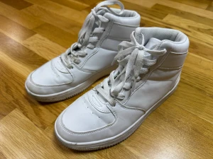 Vita höga sneakers med snörning - Snygga vita sneakers med högt skaft och klassisk snörning. Skorna har en rund tå och platt sula, tillverkade i syntetmaterial med mjukt foder. Perfekta för dig som gillar stilrena och sportiga skor med enkel design.