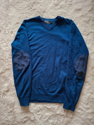 Blå stickad tröja Polo Ralph Lauren - Blå stickad tröja från Polo Ralph Lauren med v-ringad hals och förstärkta armbågslappar i mörkare blått tyg. Klassisk design med ribbade muddar och diskret logga på bröstet. Perfekt för dig som gillar stilrena och tidlösa plagg. Den passar storlek S