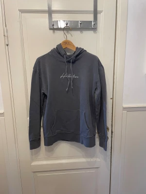 Grå hoodie från Hollister med broderi - Snygg grå hoodie från Hollister med vit broderad logga på bröstet och huvan. Tröjan har klassisk känguruficka, snörning i halsen och mjukt material som känns skönt mot huden. Perfekt för chill dagar och streetstyle. Passar xs och S