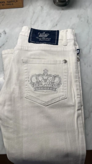Victoria Beckham vita jeans  - Rock & rebublic Snygga vita jeans från Victoria Beckham med en broderad krona och glittriga detaljer på bakfickan. Klassisk femficksmodell med silverfärgade knappar och patch i mörkblått vid linningen. Perfekta för dig som vill sticka ut med stil.