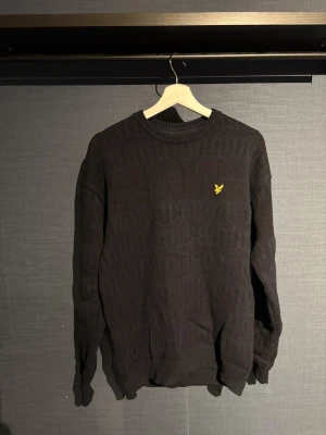 Mörkblå stickad tröja från Lyle & Scott - Mörkblå stickad tröja från Lyle & Scott med diskret mönster och gul broderad logga på bröstet. Rund hals och ribbade muddar vid ärmslut och nederkant. Perfekt för dig som gillar klassisk stil med en modern touch. Använd ett fåtal gånger