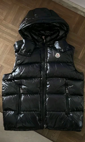 Svart dunväst från Moncler med huva - Snygg svart dunväst från Moncler med glansig finish och stor huva. Västen har två dragkedjeförsedda fickor, quiltad design och Moncler-logga på bröstet. Perfekt för kalla dagar när du vill ha både stil och värme.