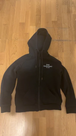 Svart Peak Performance hoodie M - Svart hoodie från Peak Performance i storlek M. Tröjan har huva med snörning, dragkedja framtill och två fickor. Klassisk logga broderad på bröstet. Perfekt för chill eller när du vill ha en enkel och stilren look.