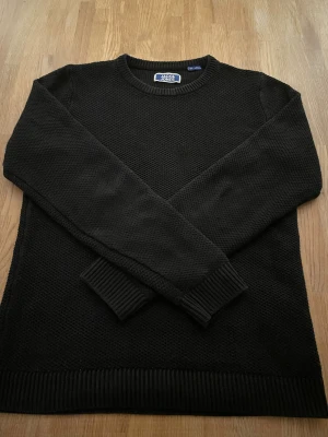 Svart stickad tröja Jack & Jones - Svart stickad tröja från Jack & Jones med rund hals och ribbade muddar vid ärmslut och nederkant. Tröjan har en klassisk, enkel design och är långärmad, perfekt för att hålla värmen under kyliga dagar. Passar dig som gillar stilrena och tidlösa plagg.