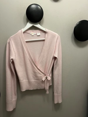 Ljusrosa omlottkofta med knyt - Supersöt ljusrosa omlottkofta från Gina Tricot med v-ringning och knyt i sidan. Koftan har långa ärmar och breda ribbade muddar vid ärmslut och nederkant. Mjuk stickad kvalitet som känns skön mot huden och ger en chill vibe.