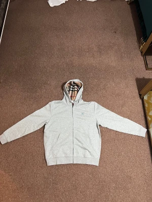 Grå Burberry hoodie med rutig huva - Säljer en grå hoodie från Burberry i storlek M. Tröjan har dragkedja, huva med klassiskt Burberry-rutigt foder och snörning. Liten broderad logga på bröstet. Mjuk bomullskänsla och två fickor framtill. Perfekt för dig som gillar stilren design med lyxig touch.
