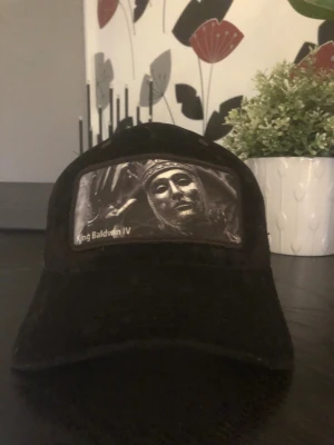 Svart King Baldwin IV keps med mesh - Säljer en svart keps med böjd skärm och mesh på baksidan för extra ventilation från hatstore. Framtill finns en patch med texten 'King Baldwin IV' och ett motiv. Justerbar snapback gör att den passar de flesta huvudstorlekar. Perfekt för dig som gillar unik streetstyle.