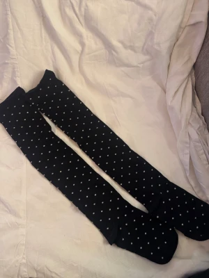 Svarta knästrumpor med vita prickar - Svarta knästrumpor med små vita prickar över hela. De är långa och går upp till knäna, perfekta för att ge lite extra stil till outfiten. Materialet känns mjukt och stretchigt, troligtvis bomull eller polyester. Prickmönstret är jämnt fördelat och ger en cool kontrast mot det svarta.