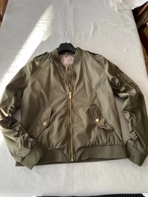 Olivgrön bomberjacka från Livelo - Snygg olivgrön bomberjacka från Livelo med guldiga detaljer och dragkedja framtill. Jackan har ribbade muddar, fickor med tryckknappar och en extra ficka med dragkedja på ärmen. Materialet är blankt och känns lätt, perfekt för vår eller höst.