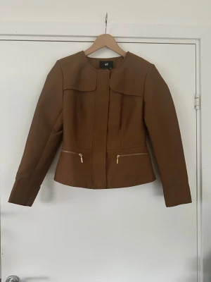 Brun kavajjacka från H&M med dragkedja - Snygg brun kavajjacka från H&M med guldfärgade dragkedjor och knappar. Jackan har en rundad krage, figurnära passform och två fickor med dragkedja framtill. Insidan har ett mönstrat foder i beige och brunt. Perfekt för dig som gillar stilrena detaljer.