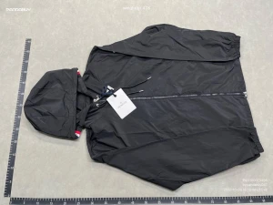 Svart vindjacka från Moncler - Svart vindjacka från Moncler med huva och snygga rödvita detaljer på insidan av huvan. Jackan har dragkedja framtill, snörning vid huvan och Moncler-logga på ärmen. Tillverkad i lätt polyester, perfekt för blåsiga dagar. Fickor med tryckknappar och stilren design. Passar för både storlek S och M.