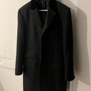 Svart covert coat med sammetskrage - Snygg svart rock/covert coat med klassisk passform och stilren design. Jackan har en sammetskrage, knappar framtill och två fickor med lock. 