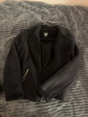 Svart teddyjacka från 915 - Svart teddyjacka från 915 med mjukt foder och krage i teddy-material. Jackan har dragkedjor vid fickorna och en stilren, loose passform. Perfekt för dig som gillar en chill och snygg look med extra komfort.