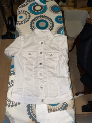 Vit jeansjacka från ONLY, storlek 34 - Snygg vit jeansjacka från ONLY med klassisk krage, silverfärgade knappar och två bröstfickor med lock. Jackan har lång ärm och figurnära passform, perfekt för dig som vill ha en fräsch och stilren look. Materialet är bomull för en skön känsla.