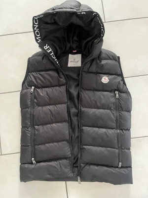Svart dunväst från Moncler - Svart dunväst från Moncler och logotyp på bröstet. Västen har två sidofickor med dragkedja och tydliga Moncler-detaljer på huvan. Endast använd 2-3 gånger. Lite stor i storlek så kan passa dig som vanligtvis har S-M. A-k0pia men i orginalutseende 