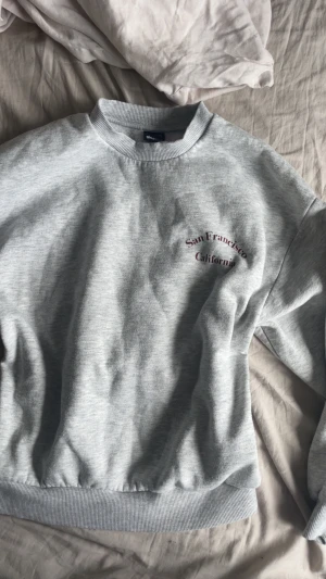 Grå sweatshirt från Ginatricot  - Grå sweatshirt från Ginatricot i storlek XS med tryck 'San Francisco California' i vinrött på bröstet. Tröjan har ribbade muddar vid hals, ärmslut och nederkant samt lång ärm. Perfekt för vintern!!💕