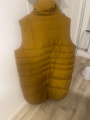 Senapsgul lång dunväst med krage - Säljer en lång, quiltad dunväst i senapsgult från Nappapi. Västen har en hög, mysig krage och praktiska fickor på sidorna. Stängs med knappar framtill och har en loose passform som ger en chill vibe. Perfekt för lager-på-lager under kyliga dagar.