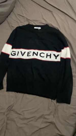 Svart stickad tröja från Givenchy - Svart stickad tröja från Givenchy med vit panel och röd kant över bröstet där loggan är broderad. Tröjan har lång ärm och rund hals, perfekt för dig som gillar exklusiva märken och stilren design.