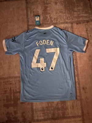 Manchester City Foden #47 matchtröja - Manchester Citys officiella matchtröja från Puma med Foden och nummer 47 på ryggen. Tröjan är ljusblå med vita detaljer, har korta ärmar och ett randigt mönster framtill. Materialet är polyester med broderat klubbmärke och tryckt sponsorlogga.
