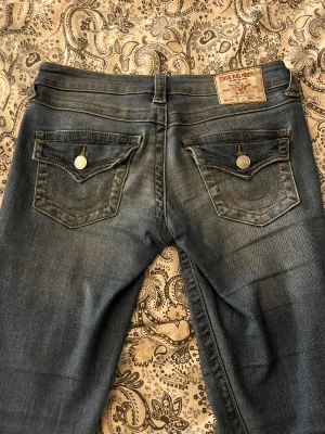 True Religion Joey Low Rise Flare Jeans - Säljer ett par blå True Religion Joey jeans med låg midja och utsvängda ben. Klassiska detaljer med markerade sömmar, stora fickor med lock och silverfärgade knappar. Jeansen är i denim och har en snygg tvättad look som ger en cool vibe. Har använt fåtal gånger så dom är i nytt skick 