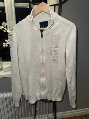 Ljusgrå kofta från Angelo Litrico - Snygg ljusgrå kofta från Angelo Litrico med dragkedja och hög krage. Tröjan har ribbade muddar och detaljerad text 'BKLYN NYC 89' broderad på bröstet. Perfekt för dig som gillar stilrena och bekväma plagg.