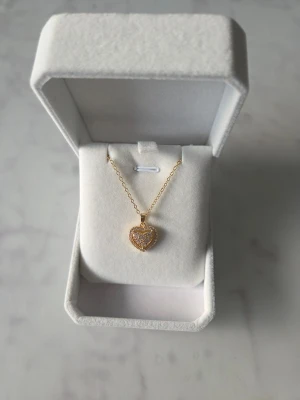 Halsband med hjärthänge - Snyggt halsband med ett hjärtformat hänge som är dekorerat med glittrande stenar. Kedjan är tunn och elegant, och hänget har en detaljrik design med små stenar som ger extra bling. Perfekt för dig som gillar romantiska smycken med lite extra sparkle.