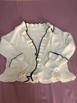 Vit kofta med volang och pärlknappar - Säljer en söt vit kofta med svarta kanter, volangdetaljer och pärlknappar framtill. Koftan har långärmade ribbade ärmslut och nederkant med volang, vilket ger en extra stylish touch. Perfekt för dig som gillar romantisk och trendig stil.💕💕