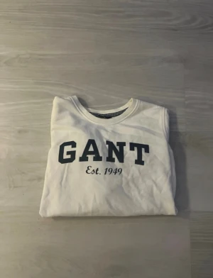 Vit sweatshirt från GANT - Vit sweatshirt från GANT med klassisk rund hals och tryck i mörkblått framtill där det står 'GANT Est. 1949'. Tröjan har långa ärmar och är gjord i mjuk bomull, perfekt för en chill och stilren look.