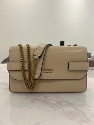 Beige axelväska från Guess - Snygg beige axelväska från Guess med guldfärgad kedja och stilren rektangulär form. Väskan har ett lock med magnetstängning, Guess-logga i guld och en praktisk dragkedja på baksidan. 