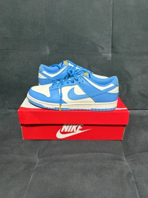 Nike Dunk Low blå och vita sneakers - Nike Dunk Low sneakers i fräsch blå och vit färg med en liten gul detalj. Rund tå och platt sula. Skorna har snörning och är tillverkade i skinn med syntetdetaljer. Perfekt för dig som gillar streetwear och vill sticka ut med en snygg färgkombo.