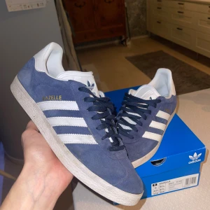 Adidas Gazelle blå mocka sneakers - Säljer ett par Adidas Gazelle sneakers i mörkblå mocka med vita detaljer och klassiska tre ränder på sidorna. Skorna har snörning, platt sula och rund tå. Adidas-logga på tungan och hälen, samt 'GAZELLE' i guld på sidan. Perfekt för dig som gillar retrostil.