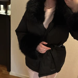 Svart pälsjacka med bälte - Säljer en svart pälsjacka med fluffiga detaljer vid krage och ärmslut. Jackan har omlottstängning med ett snyggt bälte i midjan som ger en schysst siluett. Perfekt för dig som vill ha en varm och stilren look under kalla dagar.