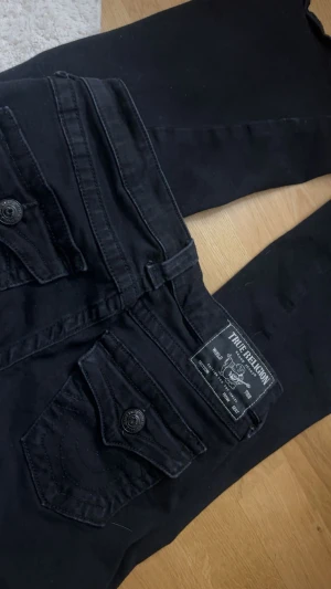 True Religion svarta bootcut jeans - Svarta jeans från True Religion, modell JOE Low Rise Flare. Snygga fickor med lock och knappar bak, klassisk logga på linningen och bootcut-ben. Jeansen har låg midja och är tillverkade i ett mjukt jeansmaterial med stretch för skön passform. Storlek 23 men dom är från kidsbrabstore så dom är lite mindre än vanliga 23