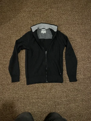 Svart kofta med dragkedja från Toms Taylor  - Svart ribbstickad kofta från Toms Taylor med hel dragkedja och ståkrage i grått. Tröjan har långärmade ärmar och breda muddar vid ärmslut och nederkant. Perfekt för dig som gillar stilren och enkel design.