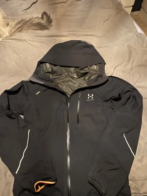 Svart Haglöfs skaljacka med huva - Säljer en svart skaljacka från Haglöfs LIM Series med huva och hel dragkedja framtill. Jackan har reflekterande detaljer på ärmarna, bröstficka med dragkedja och justerbar nederkant med orange insida. Perfekt för dig som vill ha en lätt och funktionell jacka.