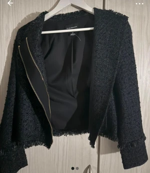 Svart kavajjacka från Club Monaco - Snygg svart kavajjacka från Club Monaco med grovstickad textur och fransade kanter. Jackan har en diskret dragkedja och är helfodrad. Perfekt för dig som gillar en stilren look med lite edge. Passar till både jeans och dressat.