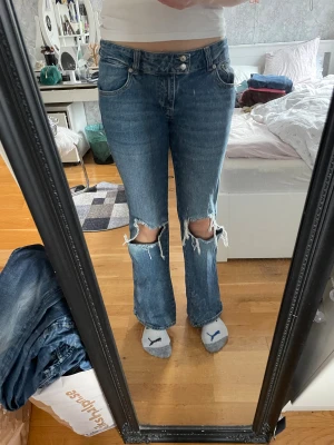 Blå ripped jeans från lager 157 - Säljer ett par blå jeans från 157 med stora slitna hål vid knäna och klassisk femficksdesign. Jeansen har raka ben och normal passform, med dubbla knappar i midjan och snygga detaljer i sömmarna. Perfekt för dig som gillar en edgy streetstil. Har råkat bleka de lite vid benen (bild 6) men det är väldigt svagt. 